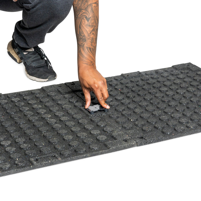 A person kneels, fitting a black connector between two Bells of Steel Rubber Flooring Gym Mat 24" x 24" tiles (Set of 2) on a white background.