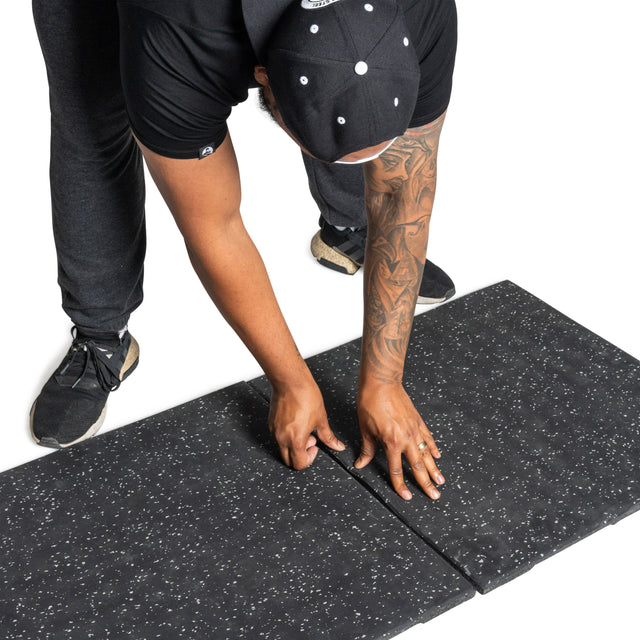 A person in black clothing and a cap installs two Bells of Steel Rubber Flooring Gym Mats 24" x 24" (Set of 2) with a speckled black design on a white background.