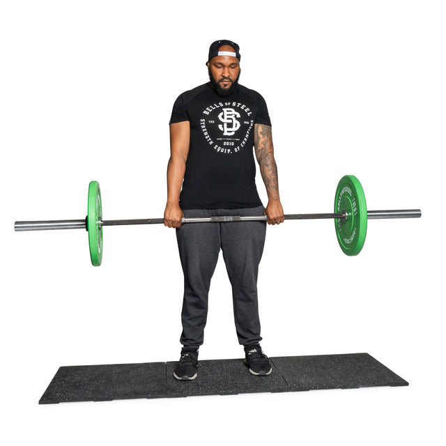 A man stands on Bells of Steel Rubber Flooring Gym Mat 24" x 24" (Set of 2), holding a barbell with green weight plates at thigh level, dressed in a black t-shirt, gray pants, and a backward cap, ready to lift.