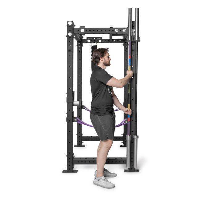 A man in a black shirt and shorts stands inside a black power rack with adjustable safety bars and multiple attachment holes, featuring Bells of Steel Hanging Barbell Holders for efficient barbell storage.