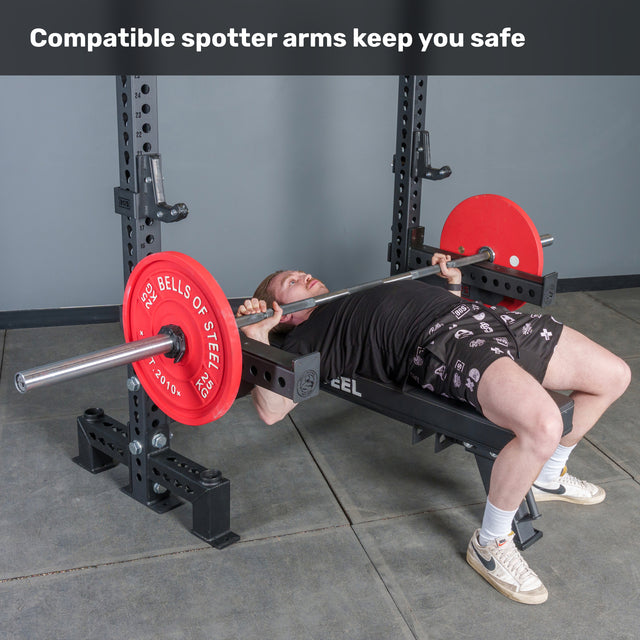 A man bench presses a barbell with red plates on the Bells of Steel Manticore Two Post Converter Bench power rack, equipped with compatible spotter arms for safety. Text at the top reads, "Compatible spotter arms keep you safe.