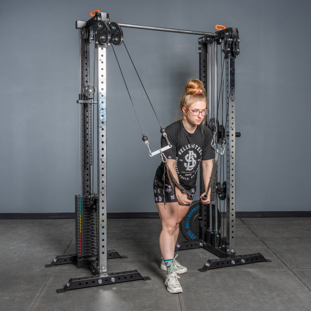 A person uses the Bells of Steel Cable Tower Squat Stands to do a cable triceps pushdown in a home gym. They wear glasses, a black t-shirt, black shorts, and white sneakers.