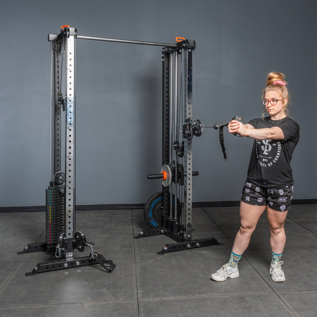A woman in glasses, a black t-shirt, and shorts uses the Bells of Steel Cable Tower Squat Stands in a gym, gripping the handle with both hands while standing on a gray floor against a plain gray wall.