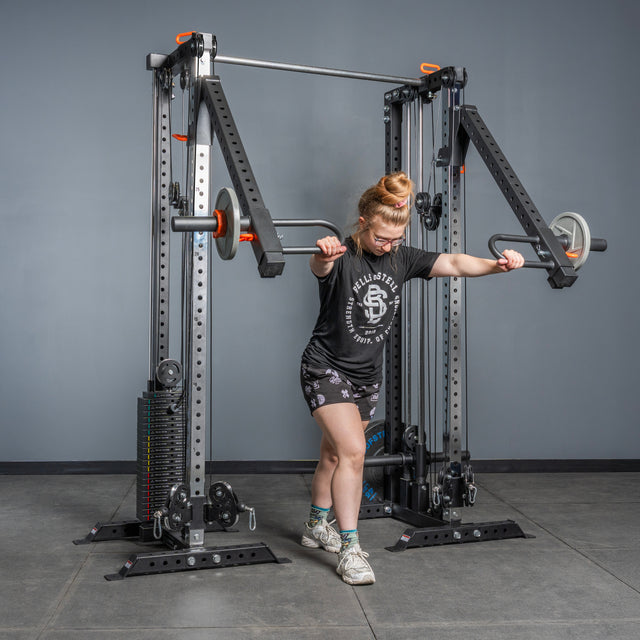 A person in athletic clothes uses the Bells of Steel Cable Tower Squat Stands, performing a standing chest press with both arms on a gym floor.