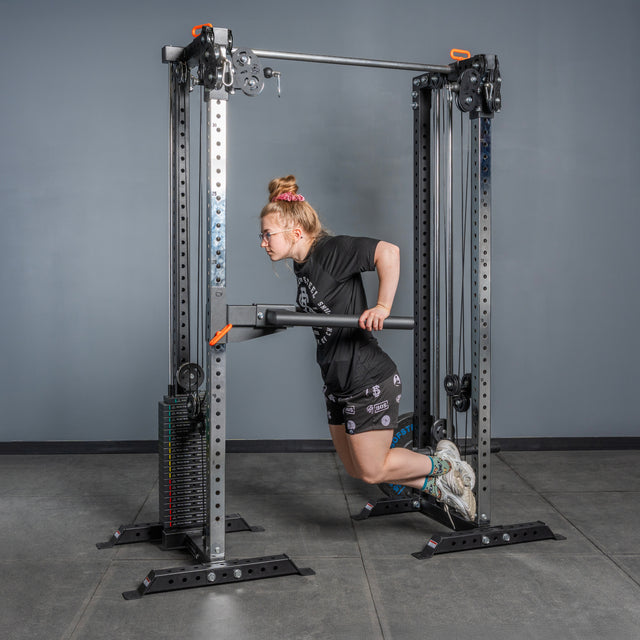 A person with long hair in a bun and glasses performs a dip on Bells of Steel Cable Tower Squat Stands at the gym, dressed in a black t-shirt, patterned shorts, and sneakers.