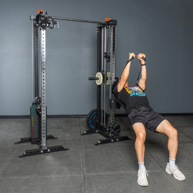 Wearing athletic clothing, a person uses the Bells of Steel Cable Tower Squat Stands in a gym with gray walls and a concrete floor, pulling the handles upward while leaning back.