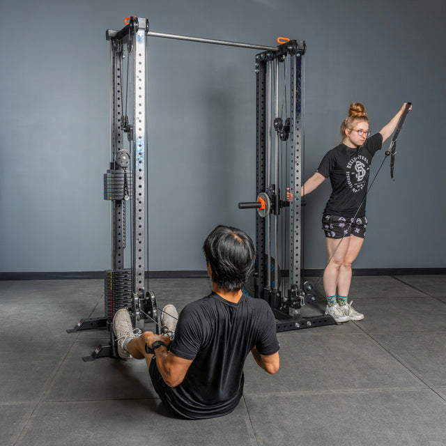 A man uses the Bells of Steel Cable Tower Squat Stands for a seated row on the floor, while a woman stands next to him with her arm extended, both working out in a modern home gym.
