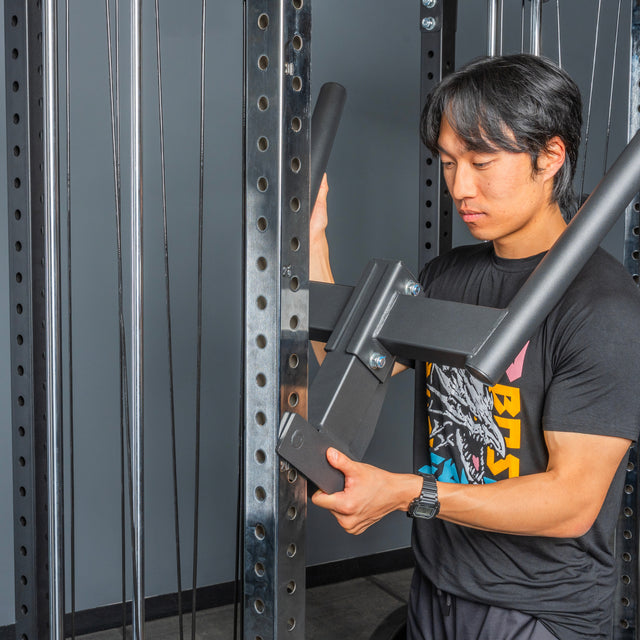 A man in a black graphic t-shirt adjusts a metal attachment on the Bells of Steel Cable Tower Squat Stands in a gym, preparing for his workout.