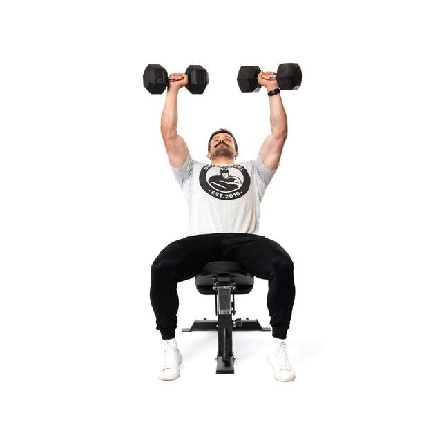 A person is seated on the Dumbbell Bench Combo by Bells of Steel, lifting dumbbells overhead. They are dressed in a gray T-shirt, black pants, and white shoes, executing a shoulder press exercise with precision against the plain white background.
