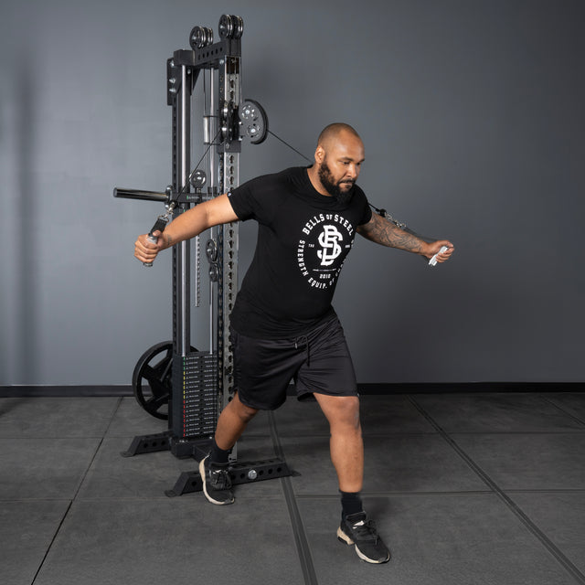A man in a black t-shirt and shorts performs a cable chest fly on the Bells of Steel Oblivyon Tower - 3" x 3” Cable Tower, standing with one leg forward and arms extended, gripping the machine handles.