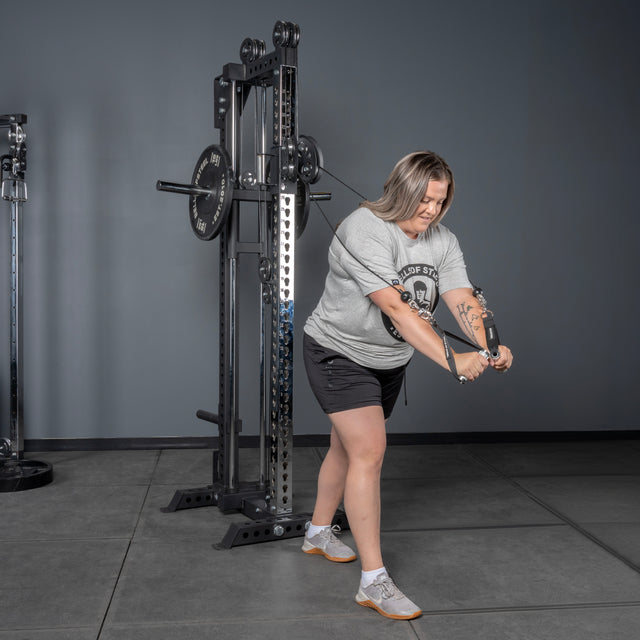 A woman in a gray t-shirt and black shorts uses the Bells of Steel Oblivyon Tower - 3" x 3” Cable Tower at the gym, performing a cable crossover by pulling the cables diagonally downward with both hands.