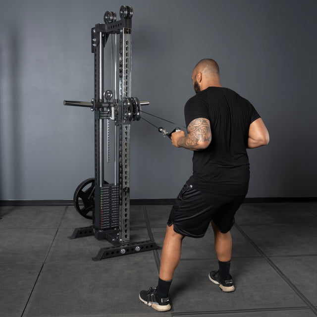 A man in black athletic wear uses the Bells of Steel Oblivyon Tower - 3" x 3” Cable Tower for strength training in a gym with gray walls, flooring, and sturdy uprights.