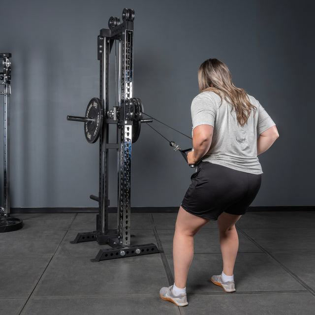 A woman in a gray shirt and black shorts uses the Bells of Steel Oblivyon Tower - 3" x 3” Cable Tower in the gym, performing a standing cable row by pulling the attachment toward her torso with both hands.