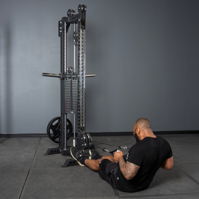A person sits on the floor in a gym, facing the Bells of Steel Oblivyon Tower - 3" x 3” Cable Tower, performing a seated row with both hands on the handles and feet braced against the uprights’ footrests.