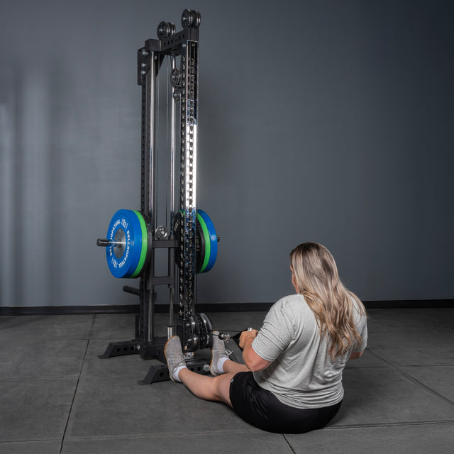 A person with long blonde hair in a gray shirt and black shorts sits on a gym floor using the Bells of Steel Oblivyon Tower - 3" x 3” Cable Tower with blue and green weight plates, against gray walls and a tiled floor.