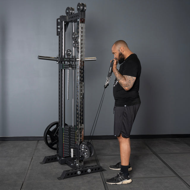 A man in athletic wear uses the Bells of Steel Oblivyon Tower - 3" x 3” Cable Tower for bicep curls in a gym with gray walls and flooring, demonstrating its home gym versatility.