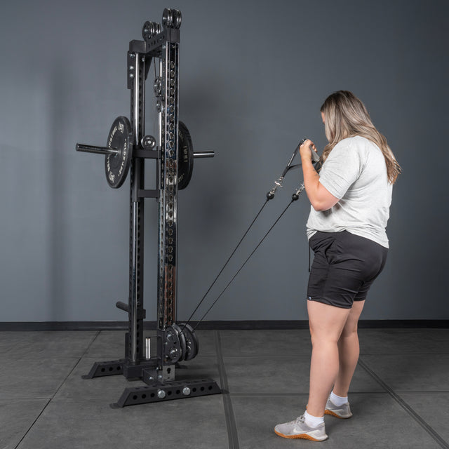 Wearing athletic clothing, a person uses the Bells of Steel Oblivyon Tower - 3" x 3” Cable Tower in a gym with plain gray walls and flooring, gripping both handles and bending their elbows while facing the machine.