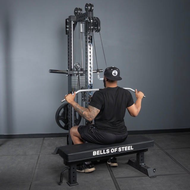 A man in a black shirt, shorts, and cap is seated on a Bells of Steel bench, using the Oblivyon Tower - 3" x 3” Cable Tower by Bells of Steel to perform a lat pulldown in a gym.