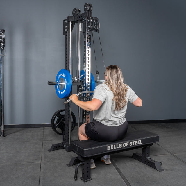 A person with long hair sits on a bench labeled “Bells of Steel,” pulling down a bar attached to the Oblivyon Tower - 3" x 3” Cable Tower by Bells of Steel in a gym setting.