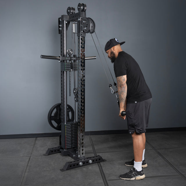 A man in black workout clothes uses the Bells of Steel Oblivyon Tower - 3" x 3” Cable Tower, standing and performing a one-arm cable pull in a gym with gray walls, rubber flooring, and sturdy uprights supporting the equipment.