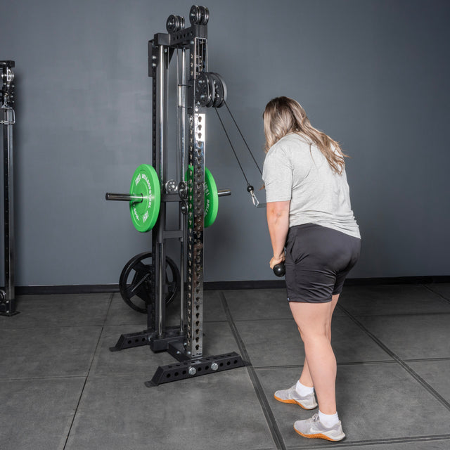 A person in a gray shirt and black shorts prepares to exercise on the Bells of Steel Oblivyon Tower - 3" x 3” Cable Tower, facing the machine and gripping the handle with both hands.