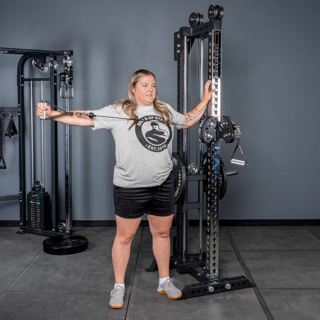 A woman in a gray t-shirt and black shorts uses the Bells of Steel Oblivyon Tower - 3" x 3” Cable Tower at the gym, performing a single-arm lateral raise with her right arm extended, standing on a gray floor against a gray wall.