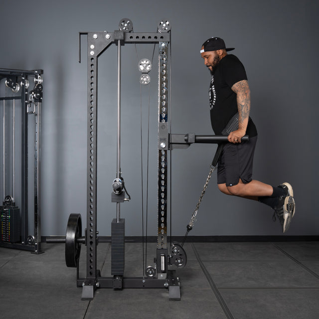 A man in a black cap, t-shirt, and shorts does a triceps dip on parallel bars attached to the Bells of Steel Oblivyon Tower - 3" x 3” Cable Tower in a gym with gray walls and flooring.