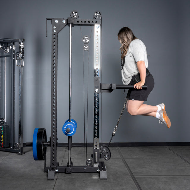 Wearing athletic wear, a woman performs an assisted dip exercise using the Bells of Steel Oblivyon Tower - 3" x 3” Cable Tower in a gym with gray walls and flooring.