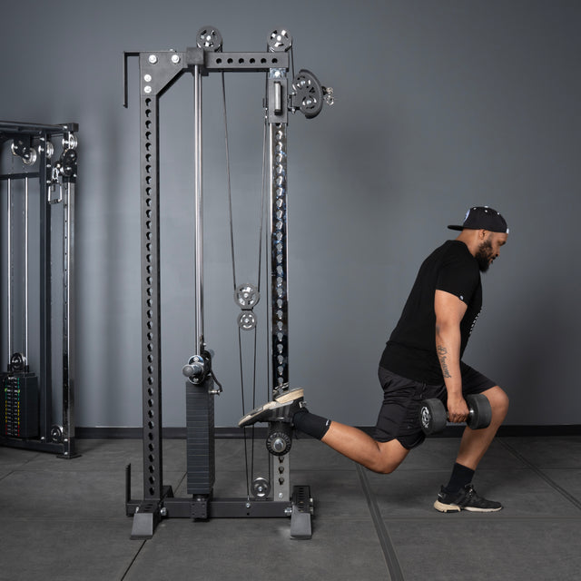 A man in athletic wear does a Bulgarian split squat holding a dumbbell, with his rear foot on the Bells of Steel Oblivyon Tower - 3" x 3” Cable Tower, inside a modern fitness center.