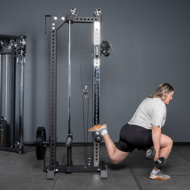 Wearing athletic clothing, a person does a Bulgarian split squat with dumbbells in a gym, using the Bells of Steel Oblivyon Tower - 3" x 3” Cable Tower for support, elevating their rear foot on the machine.