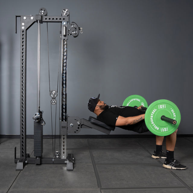 Wearing athletic gear, a person does a hip thrust on a bench, lifting a barbell with green plates in a gym featuring the Bells of Steel Oblivyon Tower - 3" x 3” Cable Tower and sturdy uprights.
