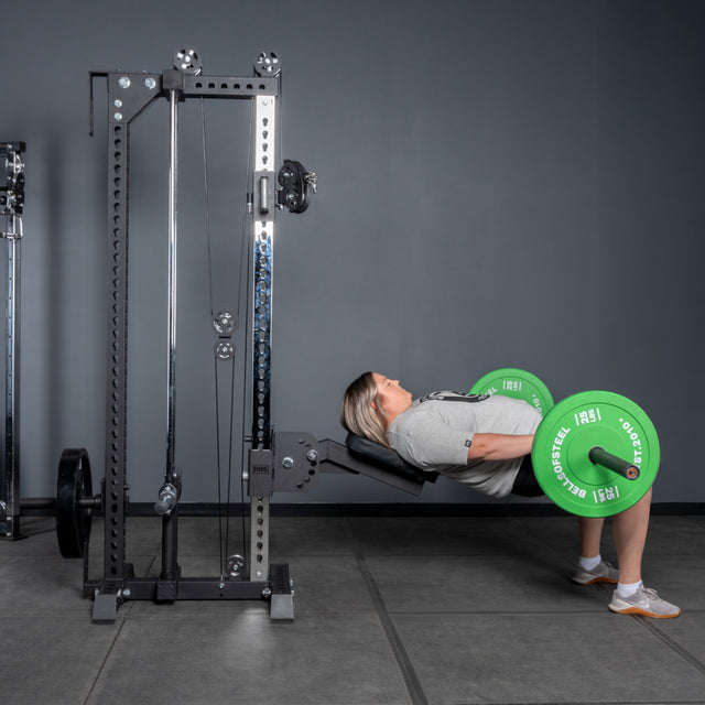 Wearing athletic clothing, a person performs a barbell hip thrust on a bench at the gym near the Bells of Steel Oblivyon Tower - 3" x 3" Cable Tower, lifting a barbell with green plates against a gray wall and floor.