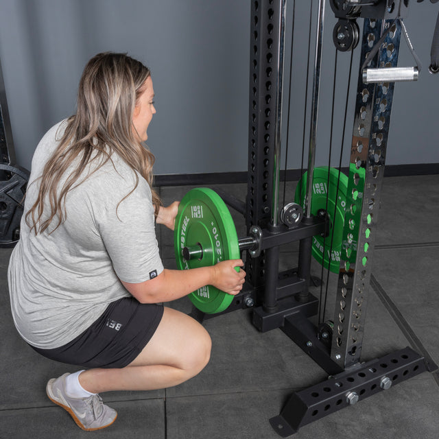 A woman in a gray t-shirt loads a green weight plate onto the Bells of Steel Oblivyon Tower - 3" x 3” Cable Tower, its sturdy metal frame perfect for any serious workout space.