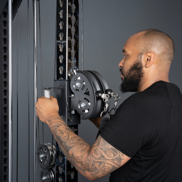 A bearded, tattooed man in a black t-shirt adjusts the weight stack of the Bells of Steel Oblivyon Tower - 3" x 3” Cable Tower at the gym, facing the equipment against a plain gray background.