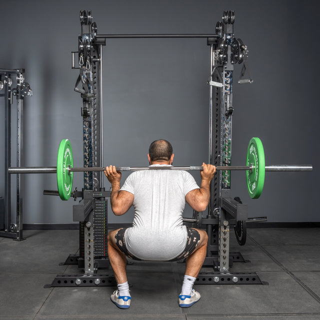 A person in a white shirt, black shorts, and white sneakers squats with a barbell on Bells of Steel Oblivyon Tower - 3" x 3” Cable Tower Squat Stands; green weight plates and an adjustable cable crossover are visible nearby.