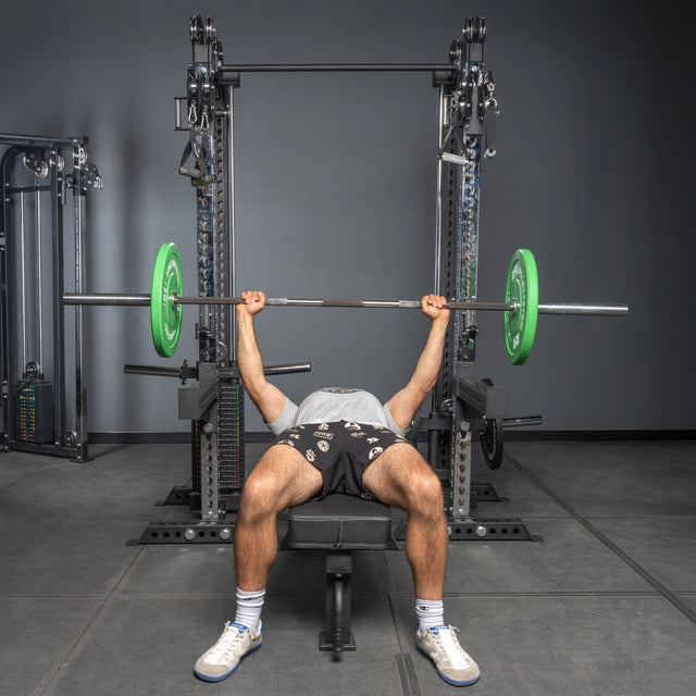 A person bench presses a barbell with green plates in a gym, using the Bells of Steel Oblivyon Tower - 3" x 3” Cable Tower Squat Stands. They wear black shorts, a gray shirt, and white sneakers.