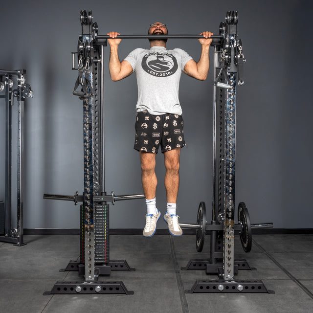 A man in a gray t-shirt and patterned shorts does a pull-up on the Bells of Steel Oblivyon Tower - 3" x 3” Cable Tower Squat Stands in a gym with gray walls and floors, lifting his chin above the bar while wearing white socks and sneakers.