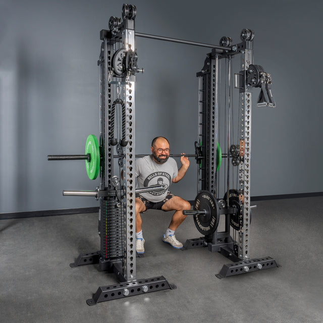 A man in a gray t-shirt and shorts performs a squat with a barbell on his shoulders using the Bells of Steel Oblivyon Tower - 3" x 3” Cable Tower Squat Stands, positioned against a gray wall.