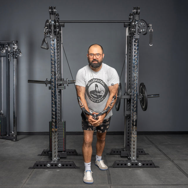 A bearded man wearing glasses, a gray Bells of Steel T-shirt, and shorts uses the Oblivyon Tower - 3" x 3” Cable Tower Squat Stands in a gym with a plain gray wall and equipment, demonstrating a cable crossover exercise.