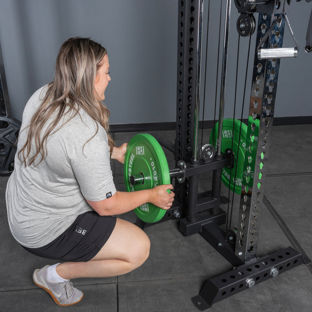 A person in a gray shirt and black shorts kneels to load a green weight plate onto a barbell on the Bells of Steel Oblivyon Tower - 3" x 3” Cable Tower Squat Stands, set in a gym with gray flooring and walls.