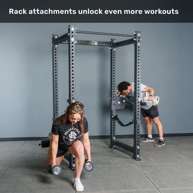 A woman performs lunges with dumbbells in front of the Bells of Steel Manticore Four Post Power Rack - Prebuilt (3" x 3", 1" Holes), while a man uses a preacher curl attachment on the rack. Text above reads, "Rack attachments unlock even more workouts.