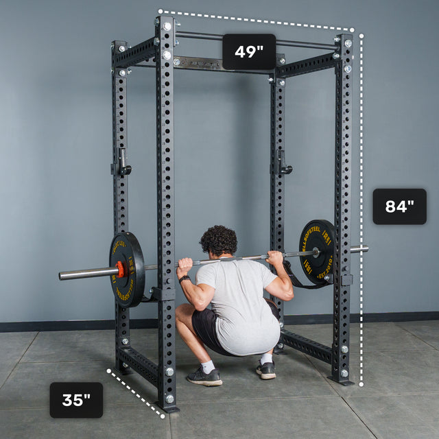 A person squats with a barbell inside a black Bells of Steel Manticore Four Post Power Rack - Prebuilt (3" x 3", 1" Holes), which measures 49 inches wide, 35 inches deep, and 84 inches tall. This sturdy power rack stands in a room with gray walls and a concrete floor.
