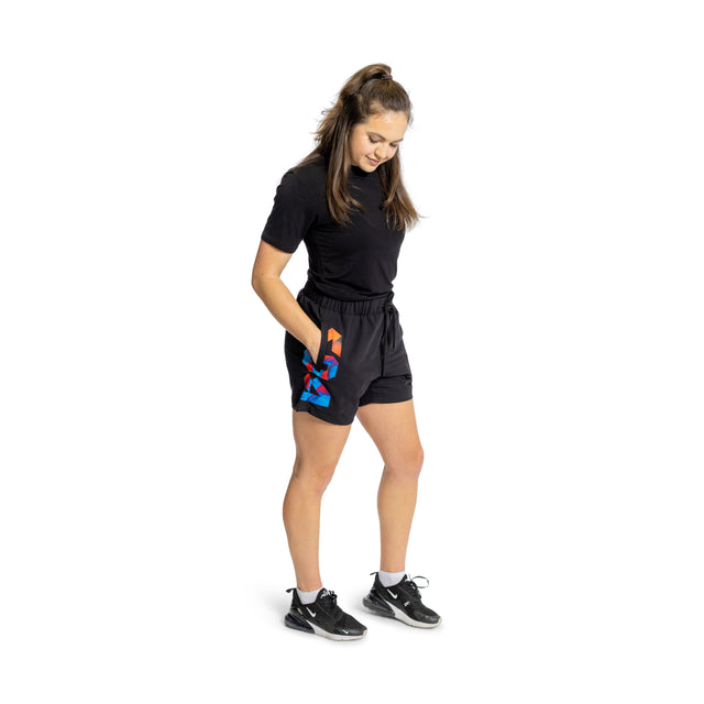 A woman smiles with hands in her pockets, wearing Bells of Steel Gym Shorts in a colorful print and a black t-shirt. Made from stretchy fabric, the outfit is paired with black athletic shoes. Her long hair is tied back against a white background.