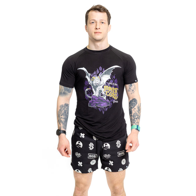 A tattooed man stands facing forward, wearing a black graphic tee and Bells of Steel Gym Shorts—black, stretchy, lightweight gym shorts with white logos—against a plain white background.