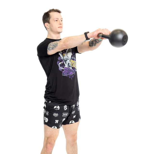A man in black graphic tee and Bells of Steel Gym Shorts performs a kettlebell swing, gripping the weight with both hands at chest height against a plain white background.