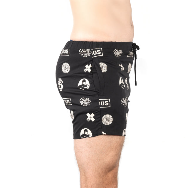 A person is shown from the waist down in profile, wearing Bells of Steel Gym Shorts with white graphic logos. These black shorts feature stretchy fabric and a drawstring waist, standing out against a plain white background.