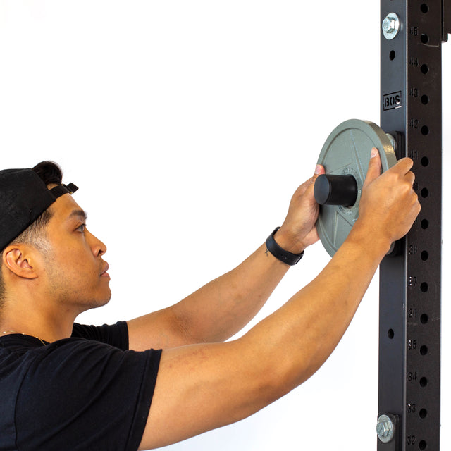 A person in a black shirt and cap loads an Olympic-sized weight plate onto Bells of Steel Bolt-On Plate Pegs attached to a power rack, against a plain white background.