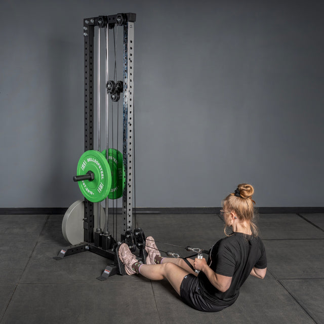 A woman performs a seated row on a Bells of Steel Footplate for Cable Tower/All In One Trainer with green weight plates in a minimalist gym with gray walls.