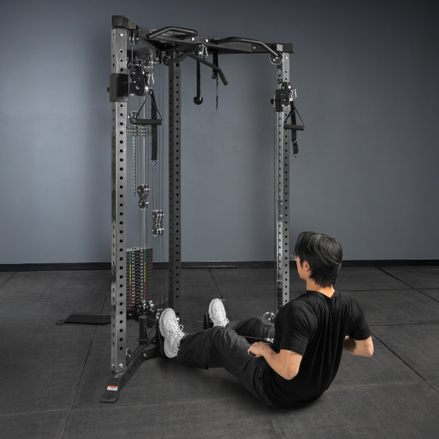 A person in black workout clothes sits on the floor, leaning back with arms behind them, facing a cable machine with a Bells of Steel Footplate for Cable Tower/All In One Trainer in a gym with gray walls and black flooring.