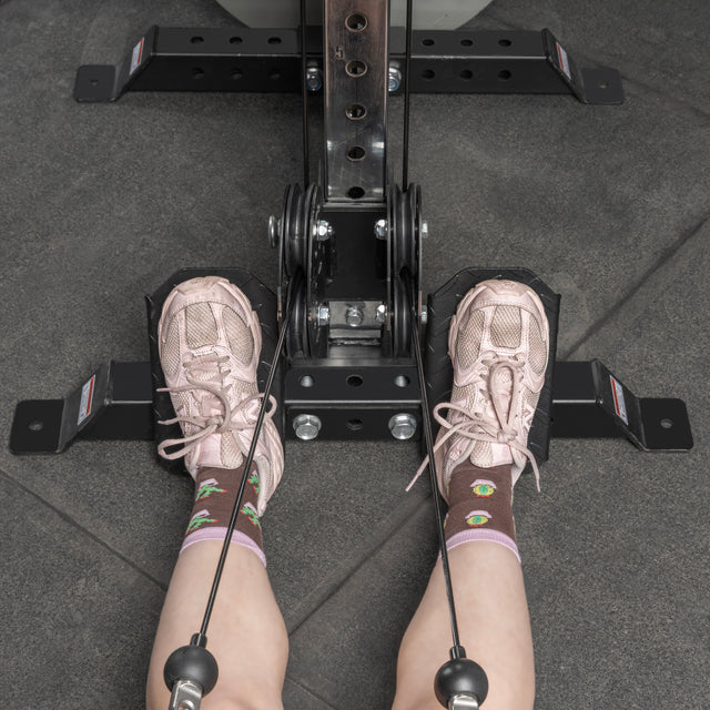 Wearing pink sneakers and patterned socks, a person sits at a rowing machine with feet on a Bells of Steel Footplate for Cable Tower/All In One Trainer, hands gripping the handles, poised to exercise atop gray gym mats.